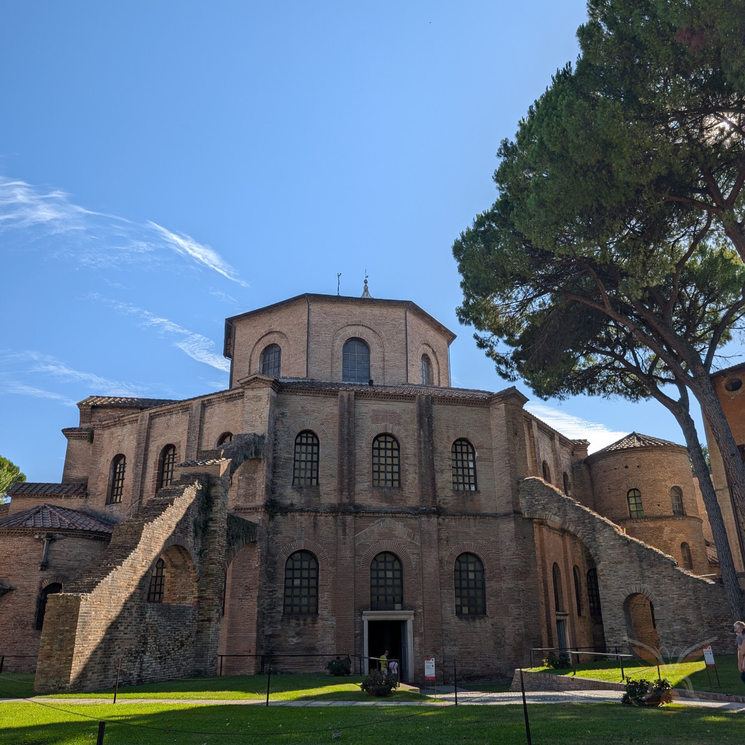 Brick exterior of the Basilica of San Vitale in Ravenna, framed by a tall pine tree.