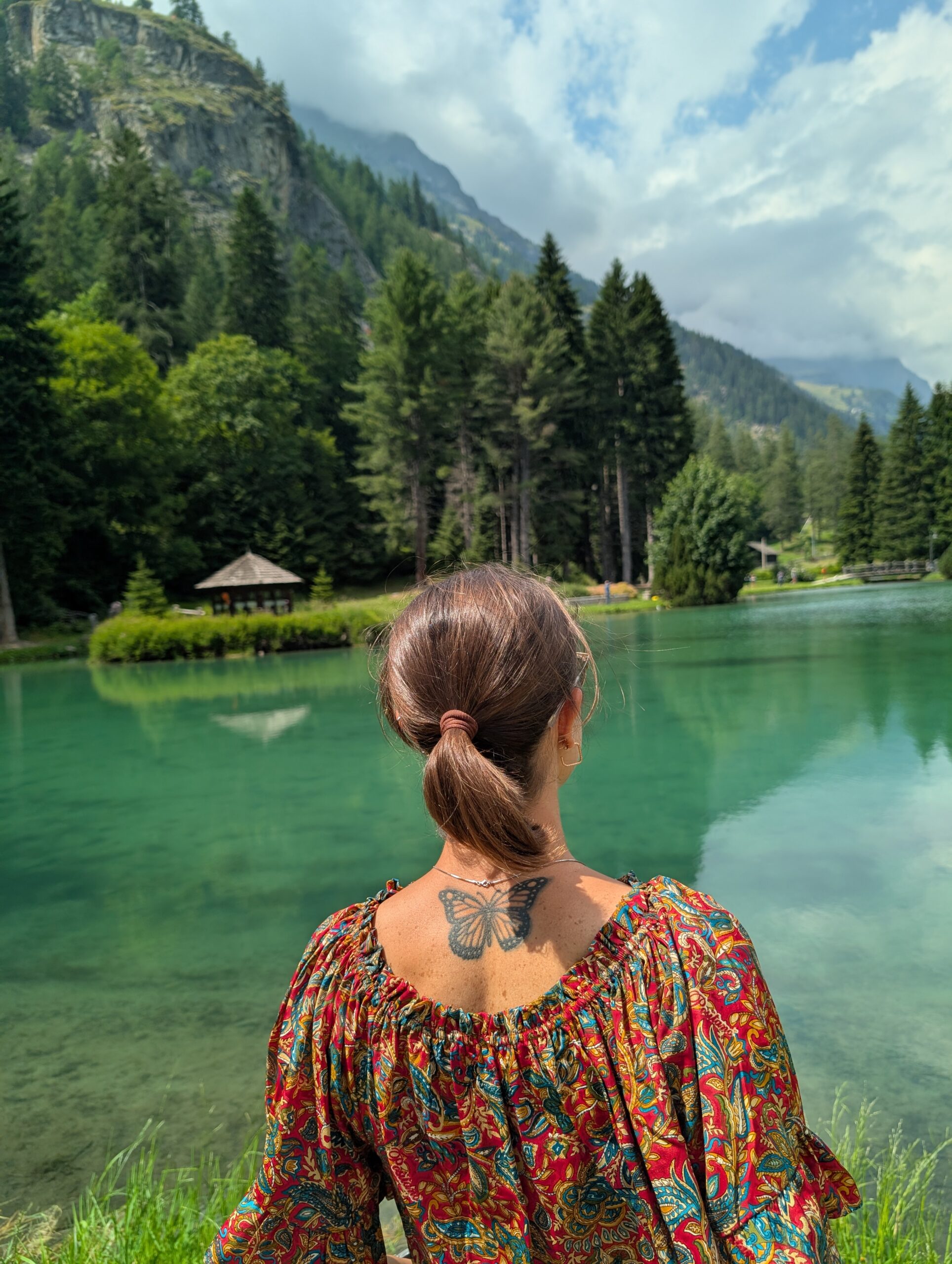 Mariachiara looking across an emerald alpine lake surrounded by forested mountains in the Aosta Valley.