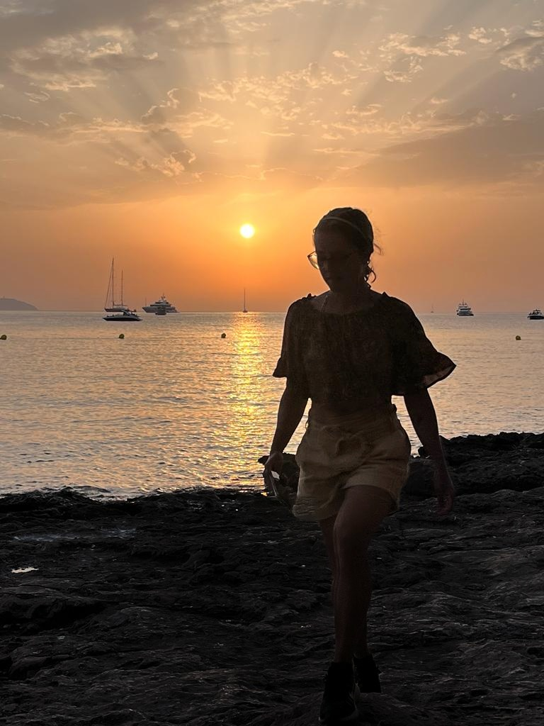 Mariachiara walking along the rocky shore at sunset in Ibiza, with sailboats on the sea.