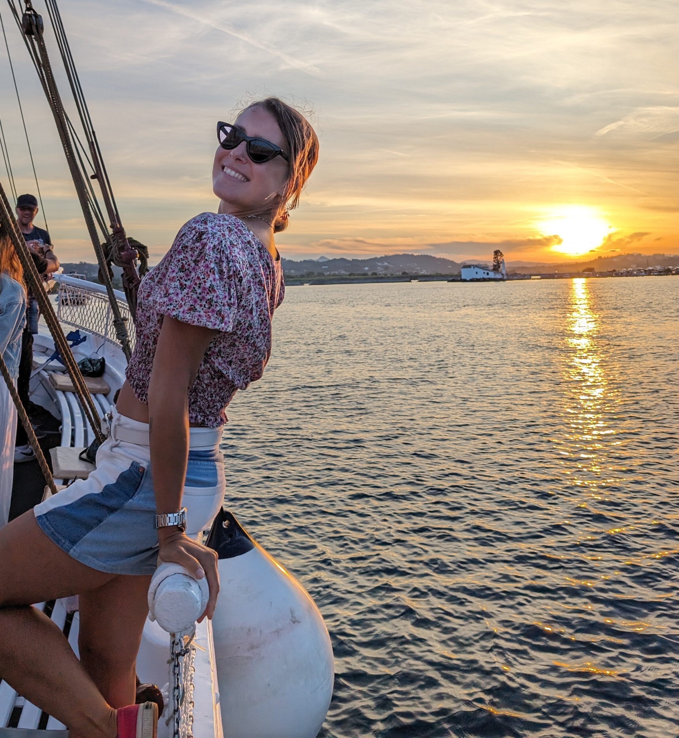Mariachiara smiling on a sailing boat at sunset, with the sea and distant hills behind her.