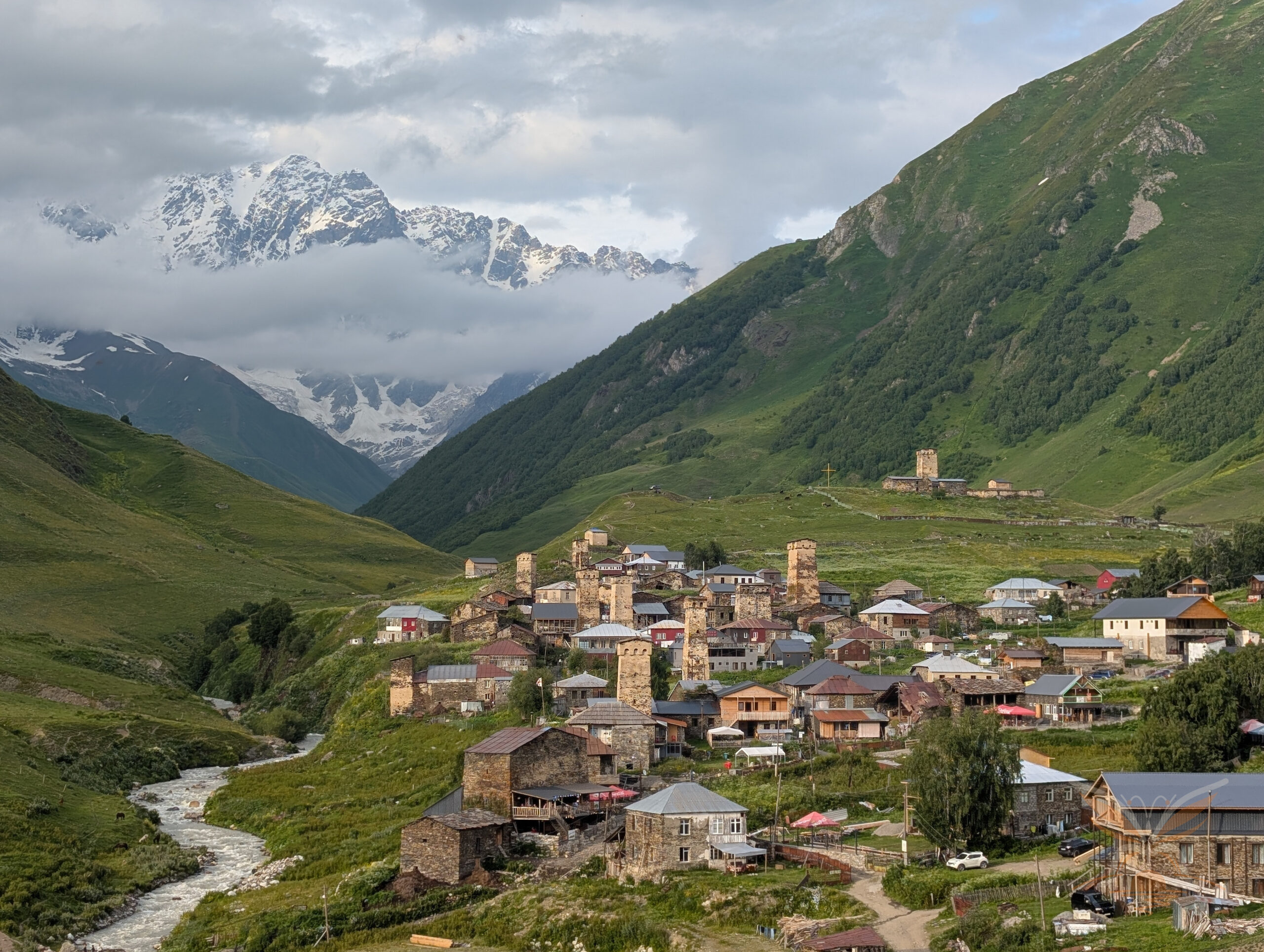 Ushguli village with Svan towers and Mount Shkhara rising through clouds behind it.