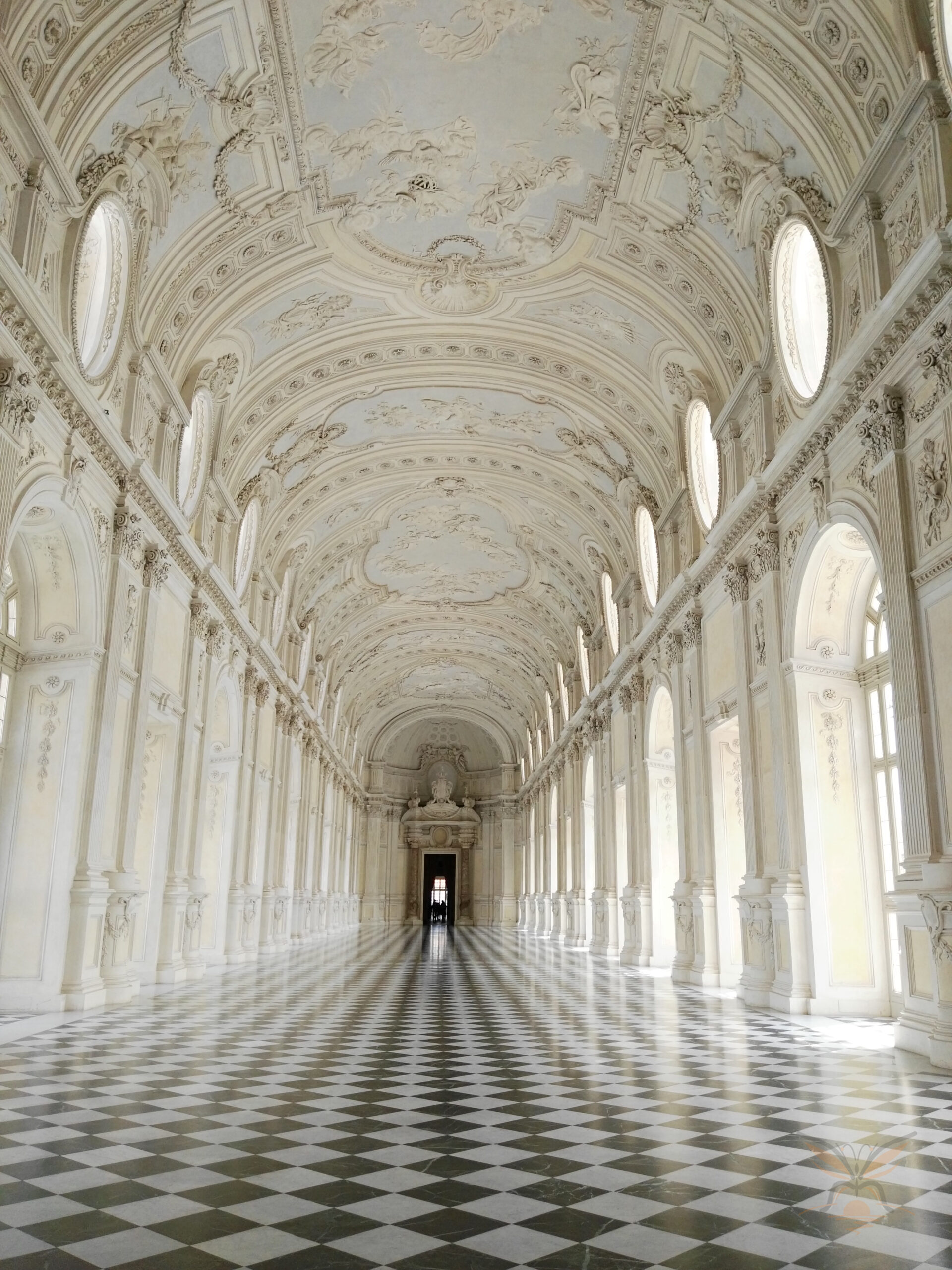 Sunlit Baroque gallery at Venaria Reale with ornate ceilings and chequered flooring.