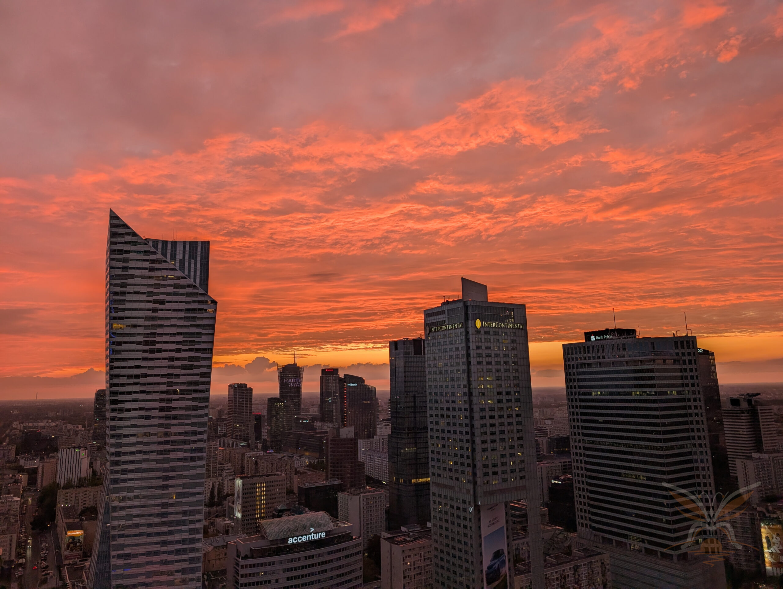 Modern Warsaw skyline beneath a dramatic red and orange sunset sky, viewed from above.